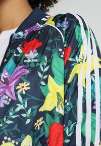 Primer plano del hombro de una chaqueta Adidas de fondo azul marino con flores rojas, amarillas y moradas, hojas verdes, rayas blancas y el logo de Adidas.