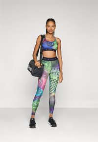 Kleurrijke sportoutfit met een gepatternde sportbeha en legging met verschillende ontwerpen. Model houdt een zwarte sporttas vast en draagt zwarte sneakers.