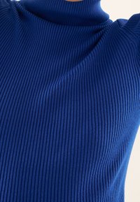 Gros plan d'un pull bleu à col roulé côtelé porté par une personne, montrant la texture et les plis du tissu sur le haut du torse.