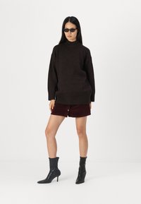 Overmaatse donkerbruine trui met een hoge hals, gecombineerd met een bordeauxrode corduroy miniskirt en zwarte haklaarsjes. Donkere zonnebril gedragen.