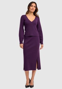 Gebreid twee-delig set in diep paars met een V-hals top en een knielange rok met zijslits. Draagt zwarte hoge hakken.