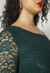 Femme portant une robe en dentelle vert foncé avec des manches transparentes, des boucles d'oreilles créoles dorées, des cheveux noirs bouclés et un rouge à lèvres rouge vif, vue du cou à l'épaule.