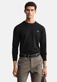 Pull noir en maille avec manches longues et col rond, associé à un pantalon à chevrons marron et une ceinture noire. Détail subtil du logo sur la poitrine.