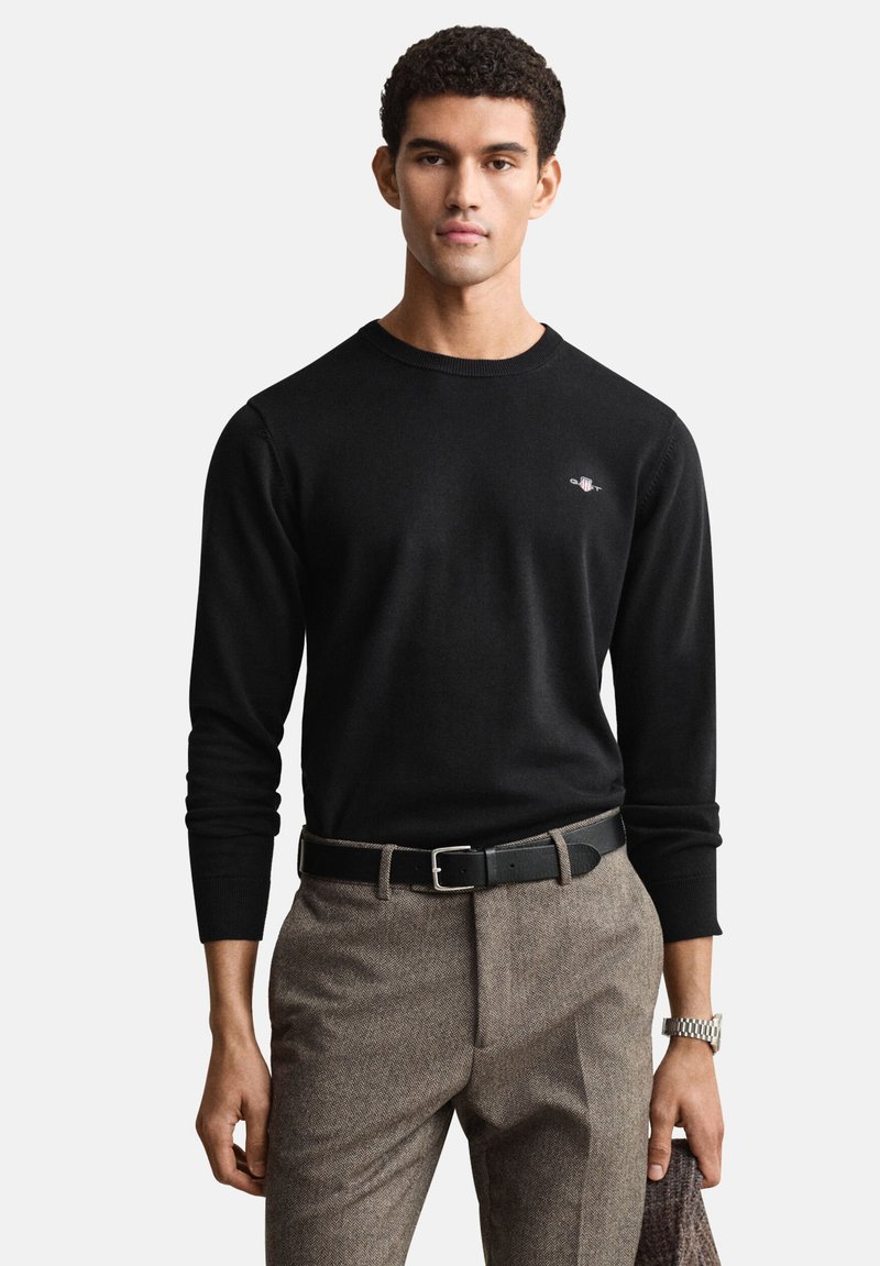 Pull noir en maille avec manches longues et col rond, associé à un pantalon à chevrons marron et une ceinture noire. Détail subtil du logo sur la poitrine.