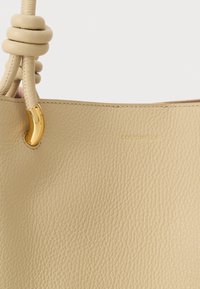 Bēša teksturēta ādas somiņa ar mezglotām virvju rokturiem un zelta metāla akcentiem, ar vieglu zīmola nosaukuma "Coccinelle" reljefu.