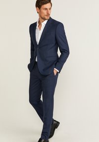 Granatowy garnitur o dopasowanym kroju, wykonany z teksturowanej tkaniny. Posiada jednorzędową marynarkę, klapy w kształcie notcha oraz slim-fit spodnie. Czarne buty.