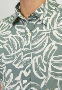 Camicia a maniche corte in tessuto verde con grandi motivi a foglia bianca, colletto con bottoni e un singolo bottone sulla patta.
