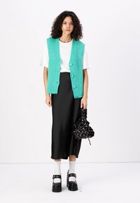 Turquoise knitted vest with buttons, worn over a white t-shirt. Black satin skirt, white socks, and platform shoes. Holding a black patterned bag.