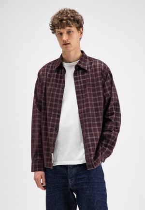 Jonge man met krullend haar die een bordeaux geruit overhemd met knopen draagt over een wit T-shirt en donkerblauwe spijkerbroek, staand tegen een effen achtergrond.