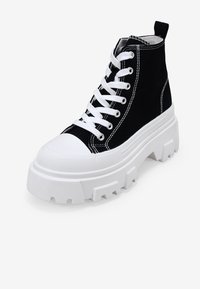 Zapatilla alta de lona negra con puntera de goma blanca y suela de plataforma. Presenta cordones blancos y costuras en contraste. Suela texturizada y gruesa.