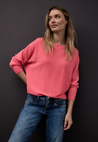 Femme aux longs cheveux blonds, vêtue d'une chemise rose à manches longues et d'un jean bleu, debout contre un mur gris foncé, regardant vers la droite.