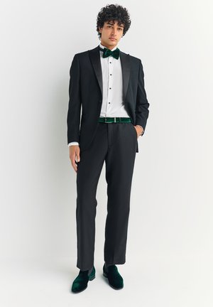 Zwarte smoking met satijnen revers, wit overhemd en groene fluwelen vlinderdas. Zwarte broek en groene fluwelen schoenen maken de formele uitstraling compleet.