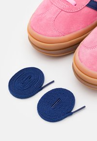 Rosa mockasneakers med bruna gummisulor och mörkblå skosnören, visade tillsammans med två rullade marinblå skosnören på en vit bakgrund.