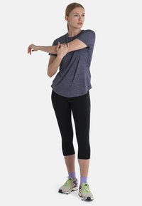 T-paita urheilullisella crew-neck-leikkauksella meriharmaana, yhdistettynä mustiin capri-leggingseihin ja monivärisiin juoksukenkään. Malli venyttää kättään.