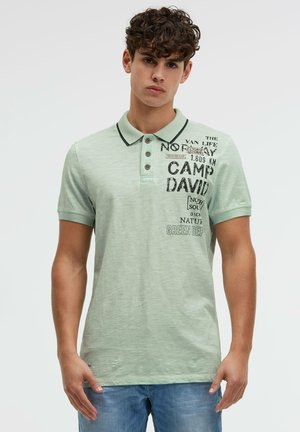 Young man wearing a light green polo shirt with black text and graphics on the chest, paired with blue jeans, standing against a white background.