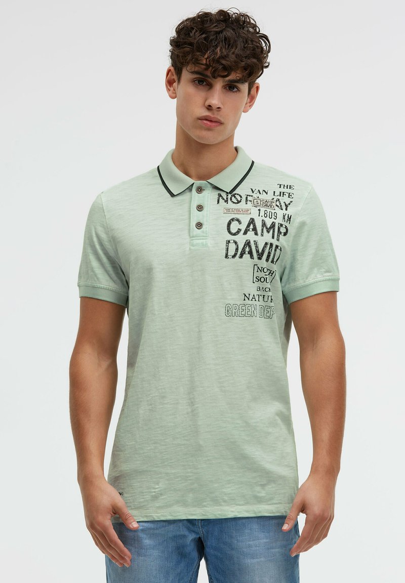 Young man wearing a light green polo shirt with black text and graphics on the chest, paired with blue jeans, standing against a white background.