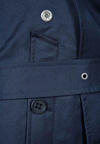 Detail tmavě modré tkaniny s knoflíkem s logem lva, pásem a kapsou, které zdůrazňují hladkou texturu a čisté švy.