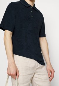 Person som bär en kortärmad svart polo och ljusbeiga byxor, håller en ljus färgad tygväska vid sin sida.