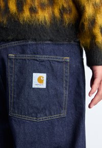 Denimbukser med en baklomme som har en oransje logo på en hvit lapp. Mørkeblå stoff med gule søm detaljer.