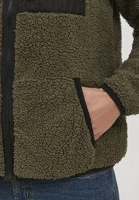 Fleece jacket with olive green texture, black accents, full zipper, and front pocket. Soft, cozy material with a contrasting panel.
