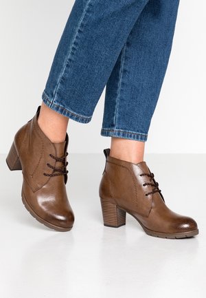 Botas de cuero marrón hasta el tobillo con un acabado texturizado, cierre con cordones y tacones de bloque, combinadas con jeans de mezclilla recortados.