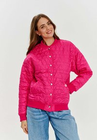 Ružová prešívaná bomber bunda s okrúhlym výstrihom, cvokmi a ribbovanými manžetami. Tkanina má hladkú textúru so geometrickým vzorom.