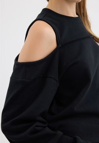 Gros plan sur une personne portant un haut noir  à épaules dénudées, exposant la partie supérieure du bras et de l'épaule.