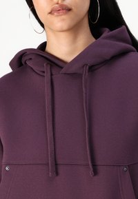 Felpa viola con cappuccio e stringa, tessuto testurizzato e tasca frontale. Include un cappuccio e un dettaglio metallico vicino alla tasca.