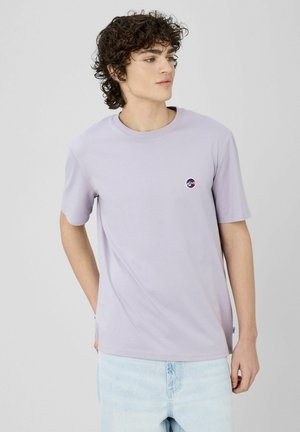 T-shirt en coton violet clair à manches courtes, col rond, avec un petit logo rond sur le côté gauche de la poitrine. Porté avec un jean en denim clair.