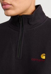 Zwarte fleece pullover met ritssluiting en hoge kraag, met een gouden logo en wijnrode tekst op de linkerkant van de borst. Soepele textuur, casual ontwerp.