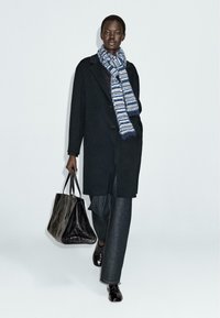 Cappotto di lana nero, sciarpa a righe blu e bianca, pantaloni in denim, scarpe di pelle nera e una borsa a tracolla nera texturizzata. In piedi contro uno sfondo semplice.
