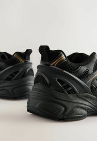 Baskets de sport noires avec tige en mesh, semelle en caoutchouc et accents jaunes. La vue latérale met en avant le design courbé et le talon texturé.