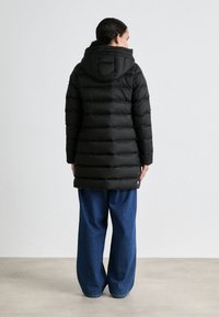 Zwarte pufferjas met capuchon, met een horizontaal gequilt ontwerp. Gecombineerd met losse blauwe denim broek. Achteraanzicht weergegeven.