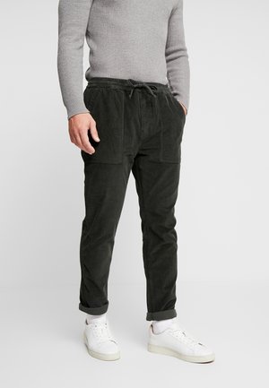 Mand iført sorte fløjlsbukser med forlommer, opkørte manchetter, hvide sneakers og en tætsiddende grå ribstrikket sweater.