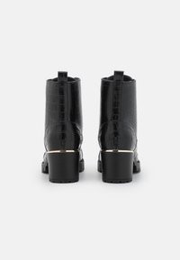 Pár černých kotníkových bot s blokovými podpatky, krokodýlí texturou a malým zlatým akcentem na podpatku, zobrazený zezadu na bílém pozadí.