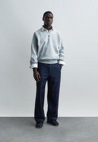 Άνδρας με ανοιχτό γκρι φούτερ πάνω από λευκό πουκάμισο με γιακά, σκοτεινό παντελόνι με ρίγες και μαύρα παπούτσια, στέκεται με το ένα χέρι στην τσέπη.