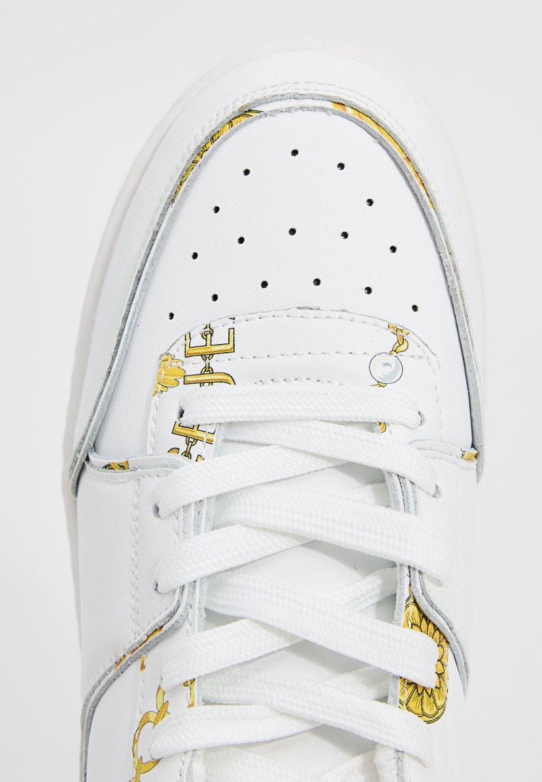 Zapatilla blanca de cuero liso con perforaciones, con acentos gráficos dorados y cordones blancos. Punta redondeada y suela de goma.
