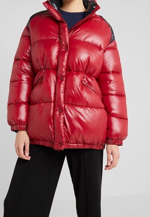 Rote glänzende Steppjacke mit schwarzen Akzenten an den Schultern, Reißverschlusstaschen und Druckknöpfen, getragen über einem marineblauen Pullover und schwarzen Hosen.