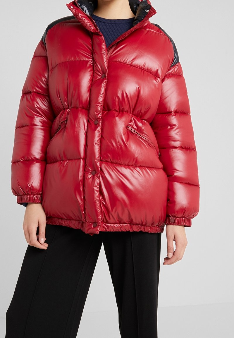 Doudoune rouge brillante avec des accents noirs sur les épaules, poches zippées, boutons-pression, portée sur un pull marine et un pantalon noir.