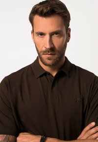 Hnědá polo košile vyrobená z bavlněné směsi, s trojicí knoflíků u límečku a embosovaným logem na hrudi. Hladký materiál, krátké rukávy.