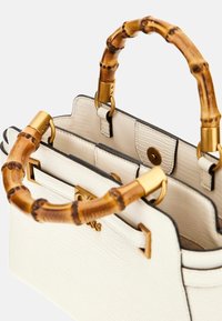 Biała torebka o teksturowanej powierzchni z bambusowymi uchwytami, złotymi akcentami oraz wewnętrznym zapięciem. Posiada przeszycia i detale na brzegach.