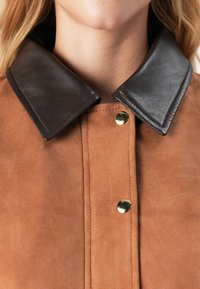 Primo piano di una giacca in suede marrone con colletto in pelle scura e bottoni a pressione dorati sul davanti, indossata da una persona.