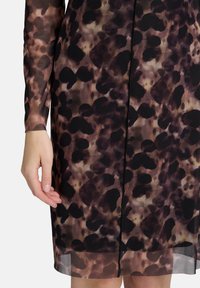 Hand und Unterarm neben einem transparenten Kleid mit schwarzen und braunen unregelmäßigen Flecken auf einem beigen Hintergrund.