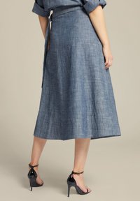 Denim wikkelrok in een middelmatige blauwe tint, knielang met een gladde textuur en strik in de taille; gecombineerd met zwarte hoge hakken sandalen.