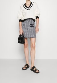 Bílý svetr s copánkovým vzorem s černými detaily, mini sukně s vysokým pasem a vzorem, s kapsami, a černé sandály se zlatými kovovými doplňky.