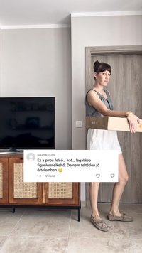 Una mujer con una blusa a cuadros y pantalones cortos blancos sostiene una caja rectangular de cartón, de pie sobre un suelo de baldosas junto a un armario de madera.