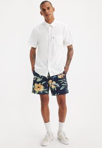 Cămașă albă cu mâneci scurte, pantaloni scurți florali navy cu modele galbene și verzi, șosete albe până la gleznă și adidași bej.