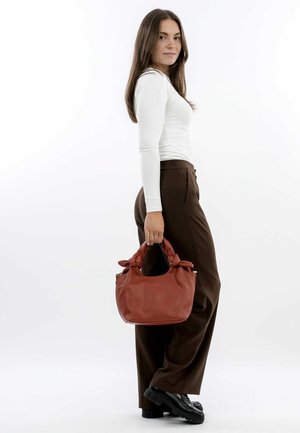 Red leather handbag with a textured surface and knotted handles. The bag has a rounded shape and a zip closure, held by a person in brown pants.