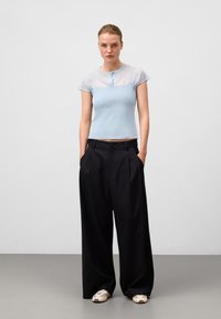 Ljust blå stickad topp med genomskinliga ärmar och en knappad halsringning, kombinerad med svarta vida byxor och avslappnade vita skor.