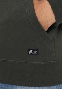 Tela de sudadera de color verde oliva, con una pequeña etiqueta azul oscuro con texto blanco. Detalle de costura visible y un bolsillo tipo canguro.